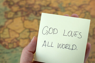 God loves all world, hand holding  handwritten inspirational quote with ancient map in the background. Close-up. Christian biblical concept of Jesus Christ's grace, mercy, forgiveness, and care.