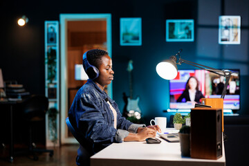 Black woman browsing the internet, and streaming movies. She attends a video call while multitasking and wearing wireless headphones. The room is bright, with a desk and a laptop on it.