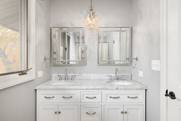 A bathroom detail with a white cabinet, chandelier hanging above the white marble countertop, and square mirrors with a reflection of the shower.