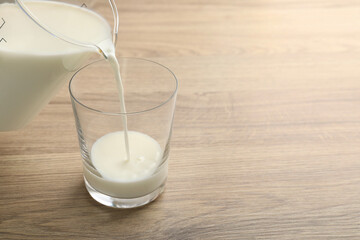 Pouring fresh milk from jug into glass at wooden table, closeup. Space for text
