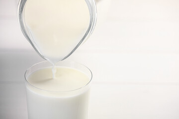 Pouring fresh milk from jug into glass at white table, closeup. Space for text