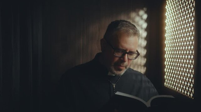 Mature priest in cassock sitting inside of confessional box and reading Bible aloud through latticed screen to parishioner in church