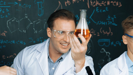 Smart teacher holding solution in beaker while student doing experiment at blackboard with chemical...