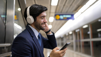 Smart business man listening music by headphone while waiting train at train station with blurring background. Skilled project manager enjoy listen relax sound while holding mobile phone. Exultant.