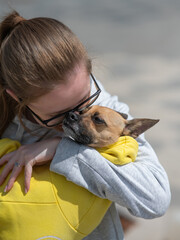 A young Caucasian woman holds a small dog in her hands in a yellow jumpsuit. Vertical photo. 
