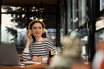 Fototapeta premium Young female student with laptop and headphones studying at table in cafe