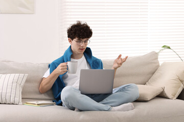 Teenager holding cup of drink working with laptop on sofa at home. Remote job