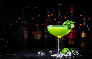Summer green cocktail drink with gin, liqueur, lime, juice, mint and ice. Black bar counter background with bottles