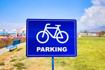 A vibrant blue traffic sign featuring a silhouette of a bicycle wheel parked against a sky backdrop with fluffy clouds. The font stands out against the natural landscape