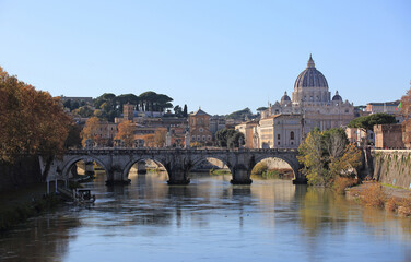 Rome, Italy - December 10, 2023: Autumnal and morning view of maple trees with Vittorio Emanuele II Bridge on Tiber River against building of St. Peter's Basilica
