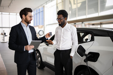 Dealer selling electric SUV to businessman in car dealership. Businessman receiving key card, dealer holding tablet. Modern electric car charging inside showroom, highlighting sustainability.