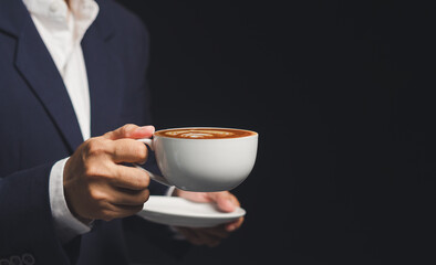 Businessman holding cup of coffee with latte art on dark background.