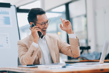 Obraz premium Excited asian male entrepreneur using smartphone and laptop, working online while sitting at workplace at home office. Businessman texting via cellphone and smiling
