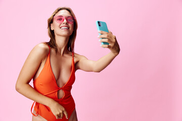 Vibrant woman in orange swimsuit capturing selfie moment on phone against pretty pink backdrop