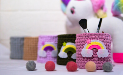 Three knitted pencil holders in pastel colors sit on a desk. Art supplies like brushes, pencils, and markers surround them, creating a playful and creative workspace