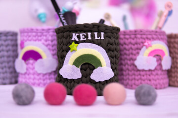 Three knitted pencil holders in pastel colors sit on a desk. Art supplies like brushes, pencils, and markers surround them, creating a playful and creative workspace
