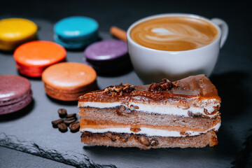 Desserts on a flat black plate. A cup of cappuccino around the sweets of colorful macaroons and carrot cake. Cinnamon sticks and coffee beans.