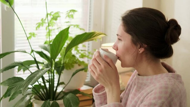 Close-up portrait of an attractive brunette woman holding a cup with her hands and taking a sip of coffee in the kitchen on a sunny morning and looking straight ahead out the window