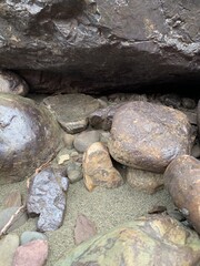 Close-up of an assortment of rocks and pebbles on a sandy beach, featuring various sizes and shapes with a smooth, polished texture. A large rock stands out among the surrounding smaller rocks.