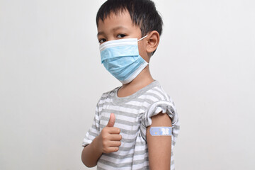 Asian boy wearing face mask giving thumbs up and showing his arm with bandage after getting vaccination. Child immunization. Protect children with vaccine concept. copy space