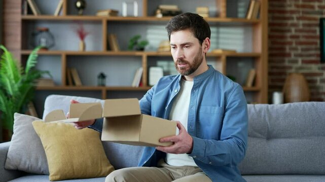 Unhappy man received a parcel with wrong product sitting on sofa in living room at home looking at camera. Bearded male is dissatisfied with shopping in online store. Service issue. Damaged item