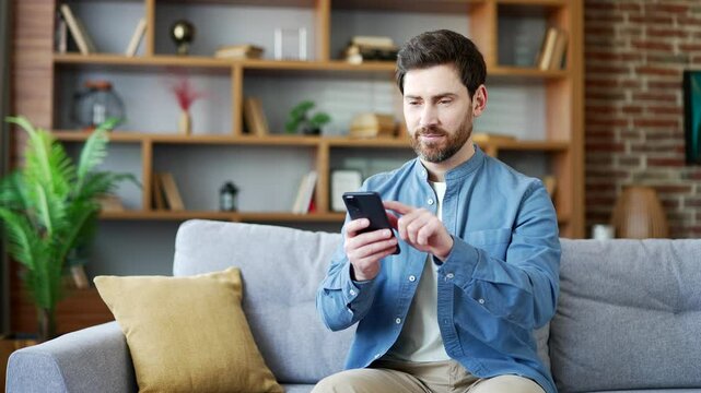 Handsome man using mobile phone sitting on sofa in living room at home. A smiling bearded male browses social networks, does shopping in an online store, chats, writes or reads messages on smartphone