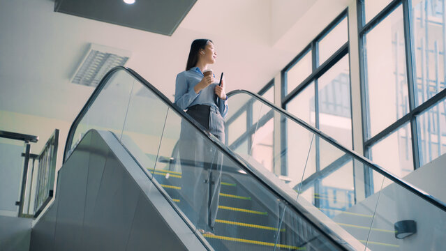 A business woman is walking down to escalator with a coffee cup in her hand - Powered by Adobe