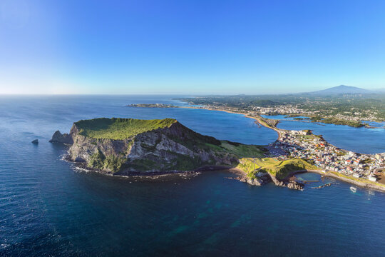 Aerial and summer view of sea and Seongsan Ilchulbong Tuff Cone with houses of Seongsanpo village against Halla Mountain near Seogwipo-si, Jeju-do, South Korea
