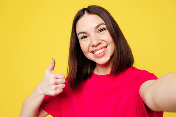 Close up young happy woman she wear pink t-shirt casual clothes doing selfie shot pov on mobile cell phone show thumb up isolated on plain yellow orange background studio portrait. Lifestyle concept.