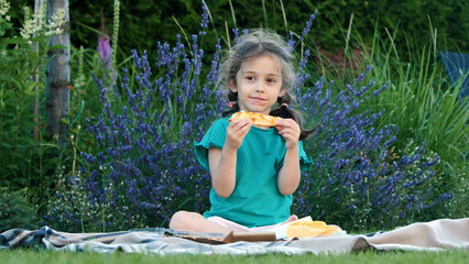 A happy girl eagerly eating a slice of pizza while sitting on a blanket at an impromptu picnic.