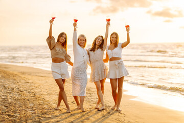 Happy women with glasses of wine embracing posing standing on the beach, celebrating bachelorette party, enjoying picnic at sunset. Vacation, relax