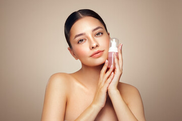 A young woman shows off a bottle with a foam dispenser for washing. Daily skin care cosmetics advertisement. Studio shoot Beige background