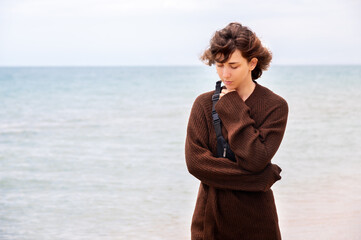 young Caucasian woman stands on the embankment by the sea and thoughtfully looks at the sand under her feet. Girl in a sweater in a pensive mood