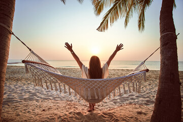 Back of woman with raised arms chilling on a hammock on a tropical island among palm trees at...
