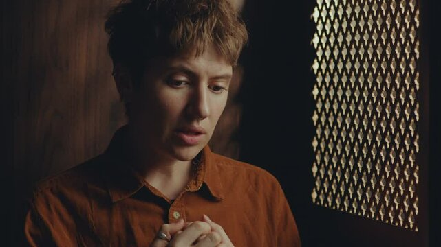 Young female penitent sitting in confessional stall and talking to priest through perforated screen when confession her sins
