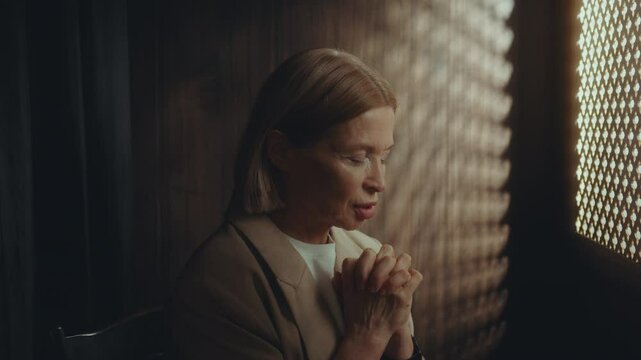Elderly woman saying prayer with clasped hands and eyes closed in dimly lit confessional booth