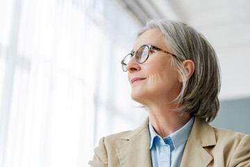 Senior businesswoman with eyeglasses smiling and looking up through office window, copy space