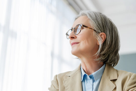 Senior businesswoman with eyeglasses smiling and looking up through office window, copy space
