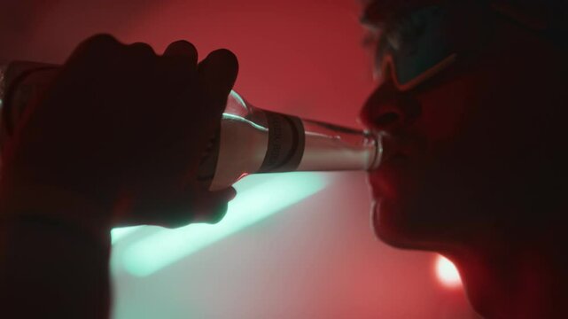Side tilt closeup of young guy in techno rave glasses holding hallucinogen drug in palm and taking it with beer while spending night in red neon lit club