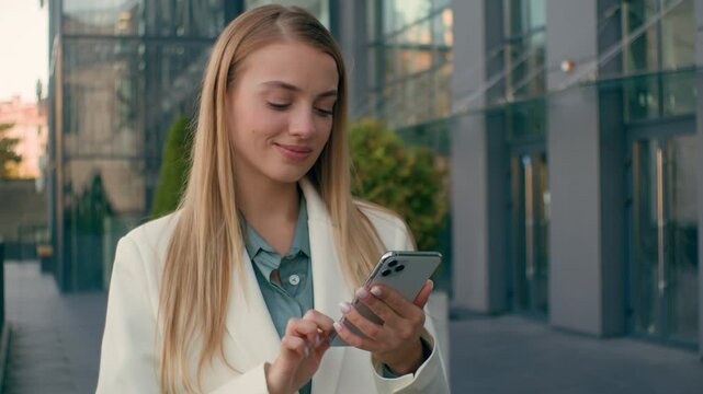 Carefree happy Caucasian woman businesswoman smile employee walk outdoors city downtown check messages business mobile smartphone phone hr manager female glad girl type scroll screen success good news
