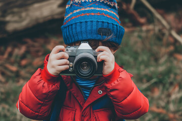 A small child, bundled up in winter attire, grips a camera and peers through its lens, taking a picture in the open air to celebrate World Photo Day.