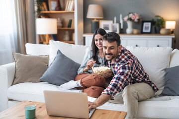 Young happy couple, simple living, sitting at home on sofa, choosing and watching movie on laptop computer, eating popcorn and enjoy time together. Healthy relationship lifestyle concept.