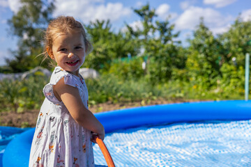 Naklejka premium A beautiful child smiles at the camera and fills an inflatable pool with water from a hose. A cute child is happy to be able to swim in the pool on hot summer days. A child fills a pool.