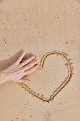 Woman's hand drawing a heart in the sand on a beach, with a heart shape in the sand next to it