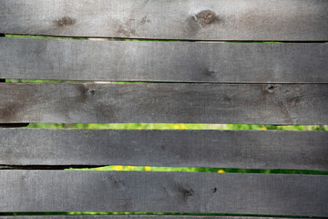 A close-up view of a weathered wooden fence with several horizontal planks. Green and yellow blur is visible through the gaps between the planks