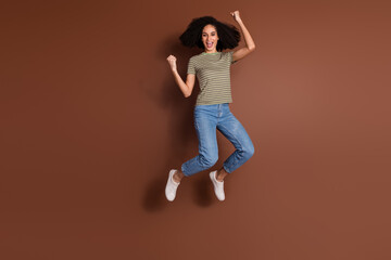 Full length photo of adorable lucky girl wear khaki t-shirt jumping high rising fists empty space isolated brown color background