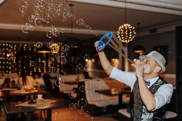 A man blowing bubbles in a restaurant. The bubbles are floating in the air and the man is wearing a hat