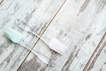 Two silicone basting brushes with transparent handles, one with a pink head and the other with a light blue head, placed on a rustic wooden white surface and copy space.