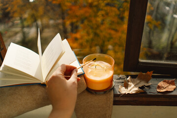 Hand with burning match lighting a candle on the windowsill with cozy autumn still life with sweater, candle and autumn decor. Autumn home decor. Cozy fall mood.
