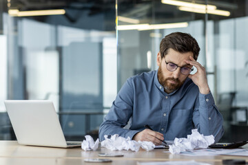 Upset and discouraged young man sitting in the office at the desk, holding his head with his hand,...